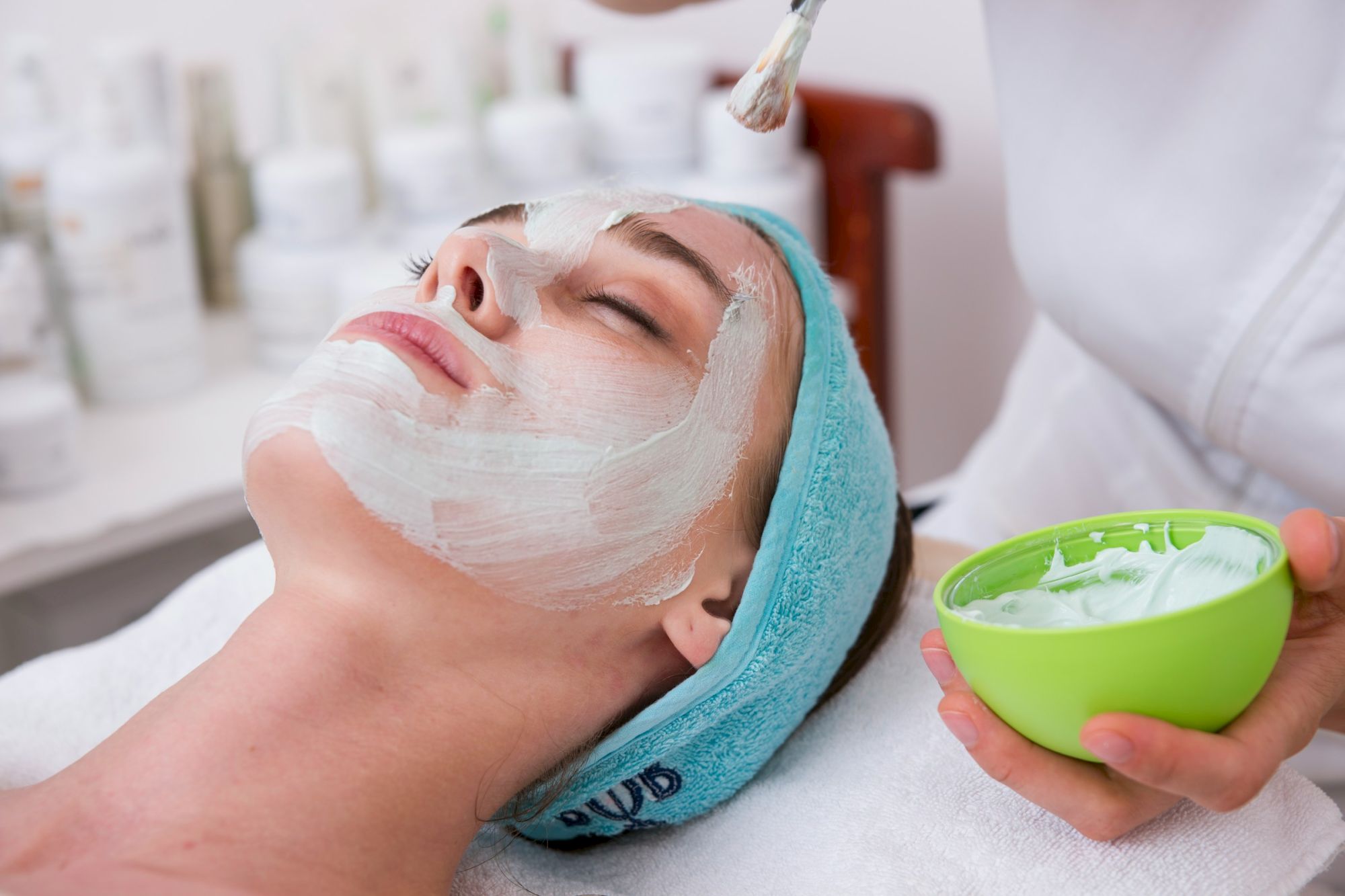 A woman receives a facial treatment: eyes closed, mask applied, head wrapped in a turquoise towel, esthetician holding a green bowl of product.