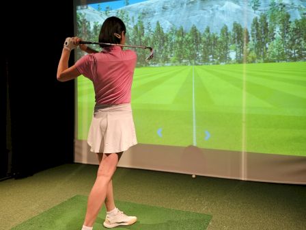 A person stands on indoor golf turf using a club to hit a ball at a projected virtual course on a big screen.
