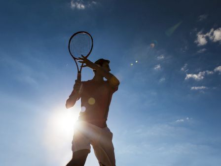A tennis player swings a racket outdoors under a bright sun and blue sky.