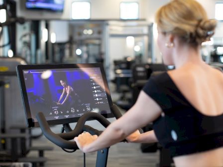 A woman pedals a stationary bike while watching fitness metrics on a screen.