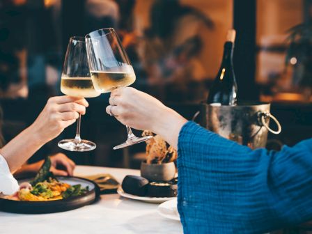 Friends clink stemmed glasses of sparkling drinks at a cozy restaurant table, toasting and chatting together.