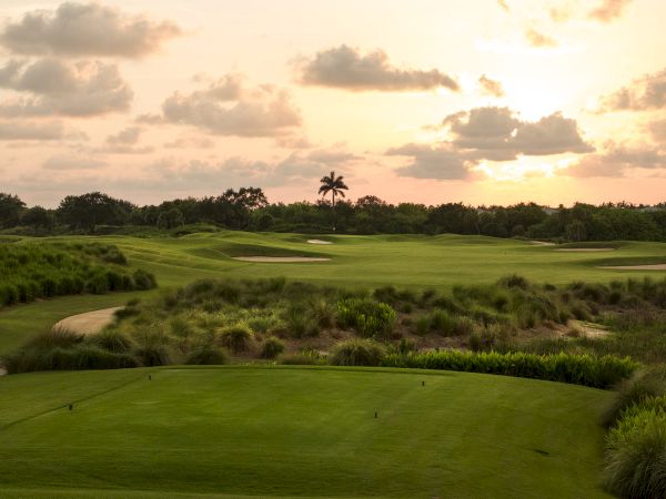 A lush green golf course with sand traps and a winding fairway, silhouetted by a warm sunset sky and clouds.