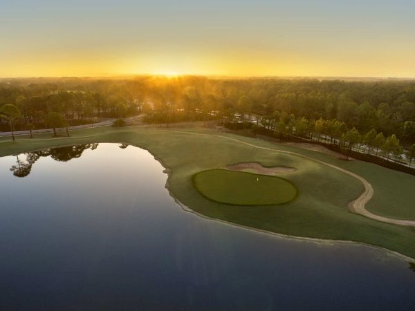 An aerial view of a golf course at sunset, with a pond, manicured greens, and trees glowing in golden light.
