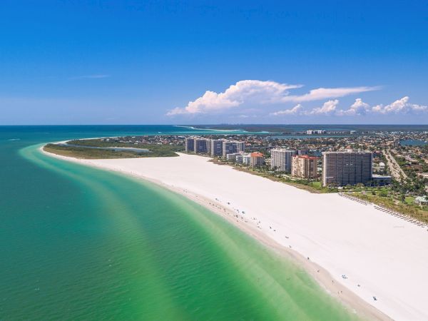 Aerial view of a wide sandy beach with ocean water, beachfront high-rises, and scattered people under a blue sky.