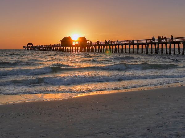 A sunset over the ocean with waves rolling in and people walking along a long wooden pier, glowing in the golden light.