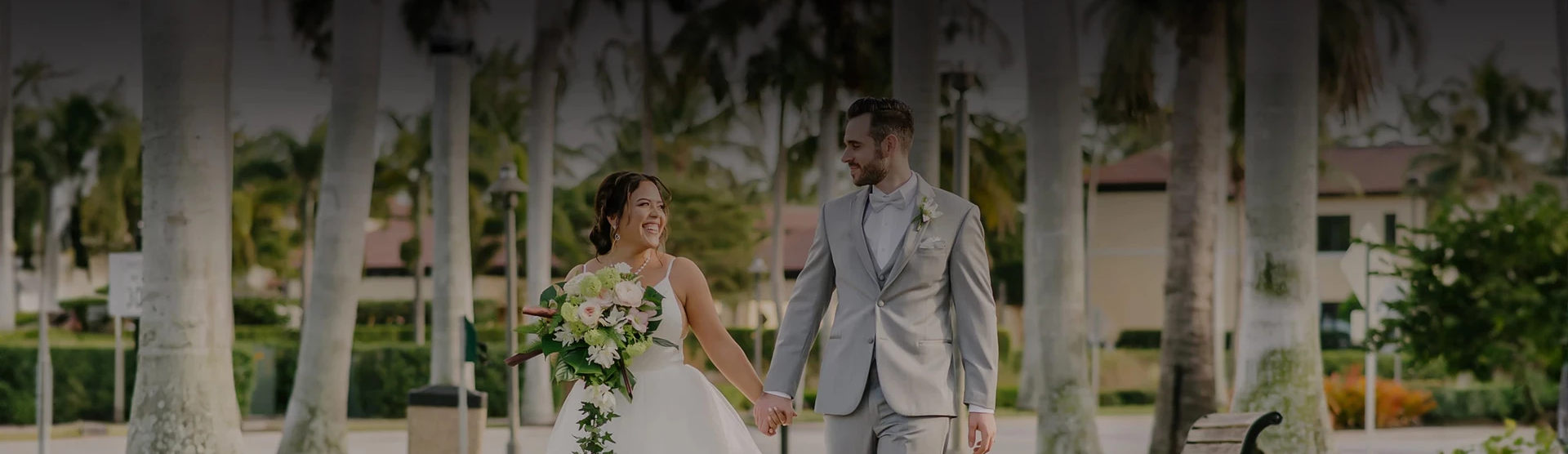 A bride and groom walk hand-in-hand outdoors, smiling, with the bride holding a bouquet, under palm trees on a sunny day.