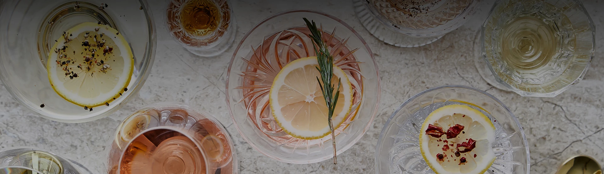 A top-down table of assorted cocktails with lemon slices, herbs, and colorful garnishes in elegant glassware, all arranged on a light surface.