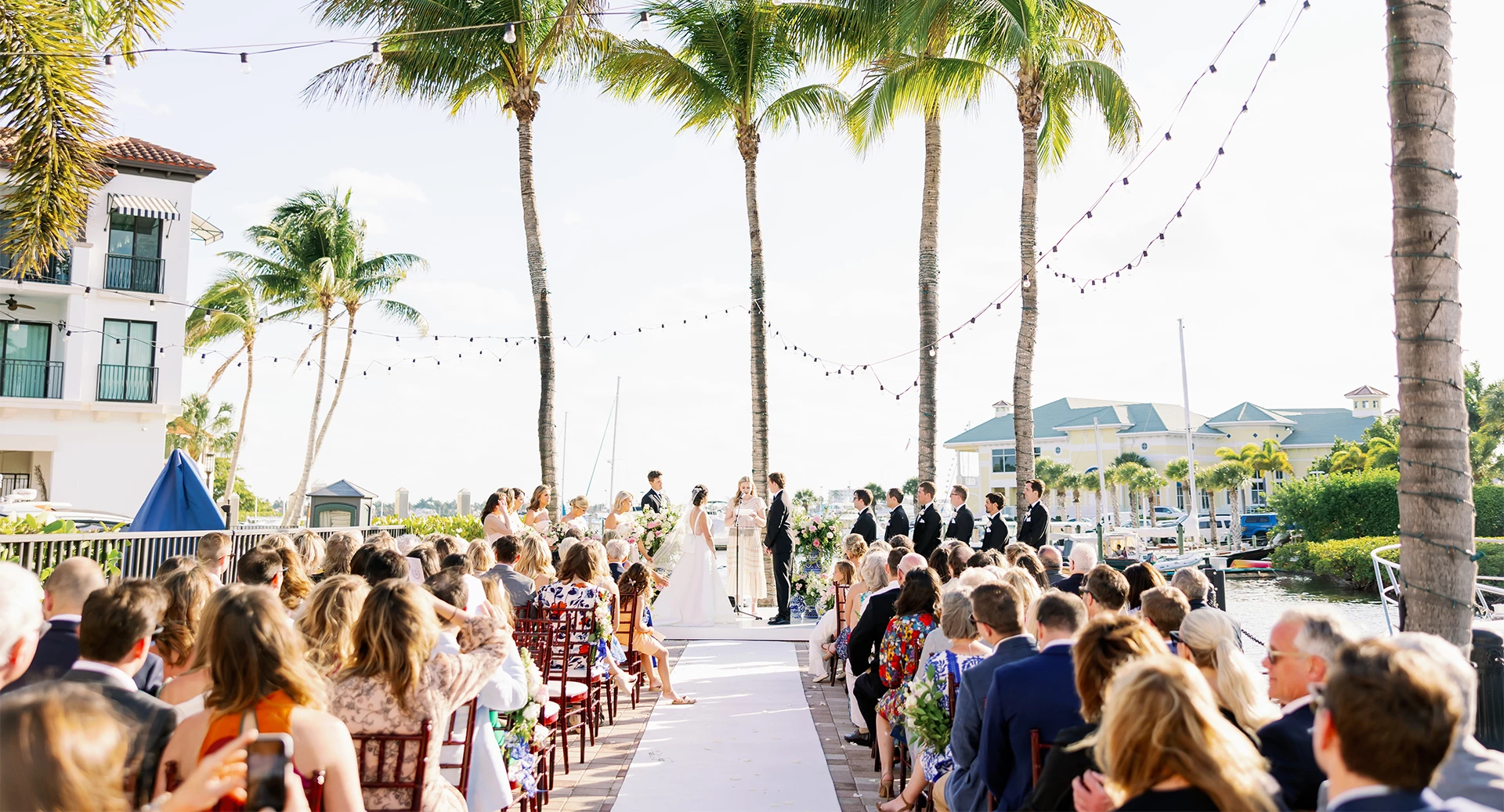 A beachside wedding ceremony with guests seated along a white aisle, tropical palm trees, string lights, and a bright seaside backdrop.