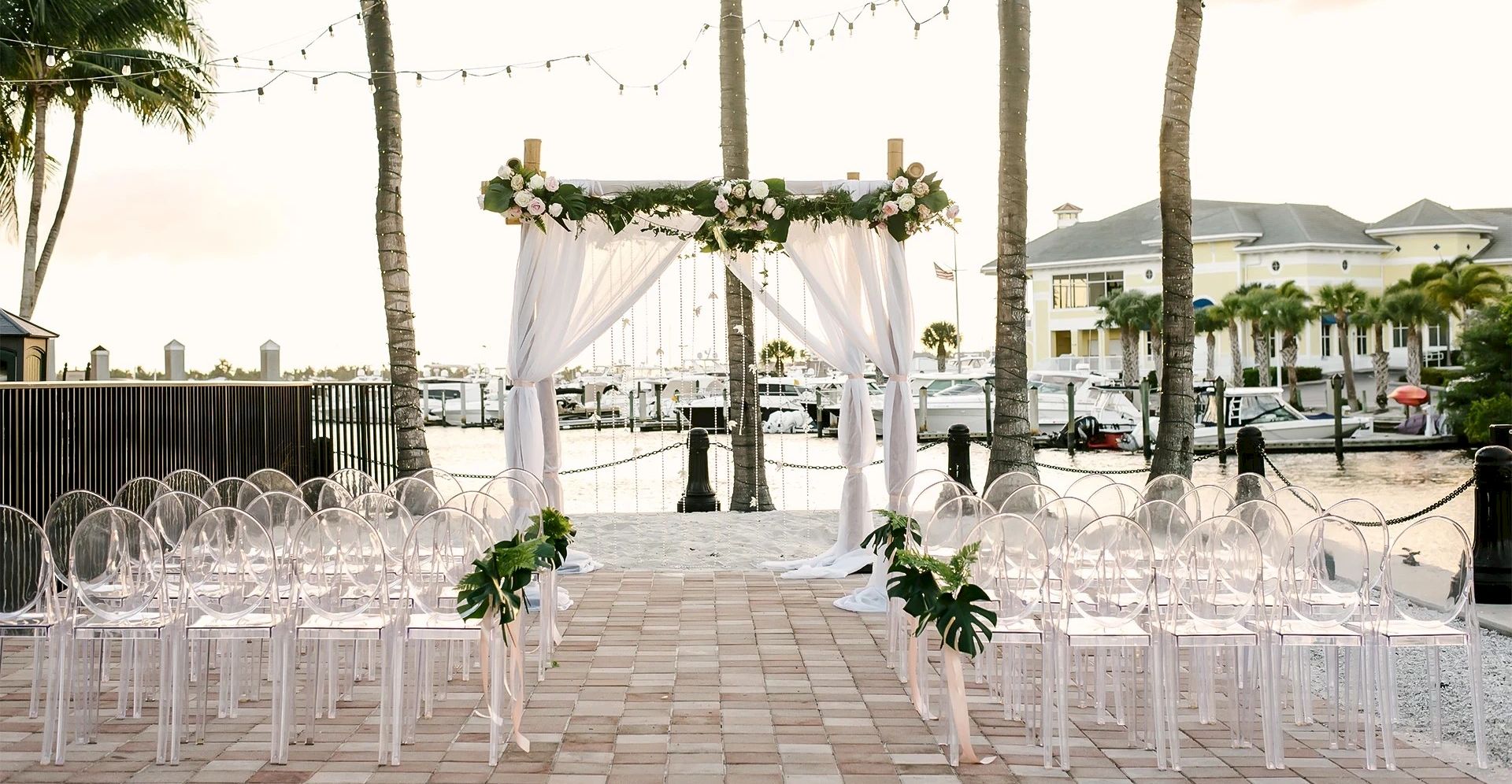 An outdoor waterfront wedding ceremony setup with white draped arch, floral decor, string lights, and clear round-back chairs.