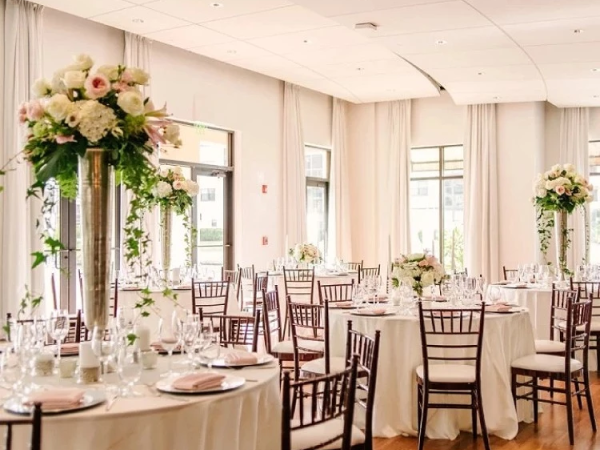 Elegant banquet hall set for a formal event with round tables, white linens, floral centerpieces, and wooden chairs, soft natural light.
