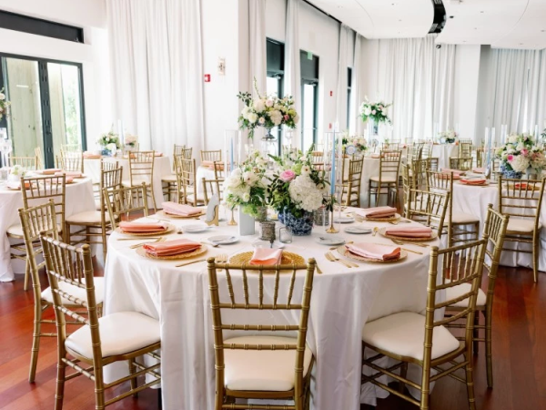 Elegant banquet setup with round tables, white linens, gold chiavari chairs, and lush flower centerpieces for a formal event.
