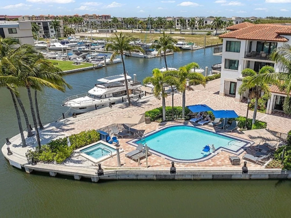 A waterfront marina view with a blue pool and hot tub, palm trees, boats, and modern condo buildings by the water, sunny day.