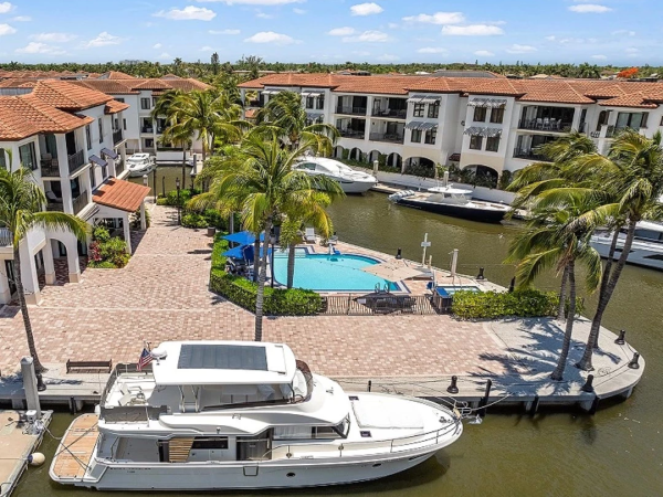 A canal-side marina with yachts, palm trees, red-tiled buildings, and a pool area between white apartment complexes along the water.