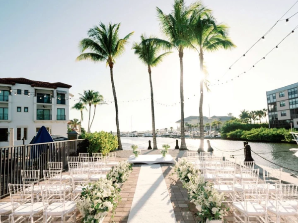 A sunny outdoor wedding setup with white chairs, floral aisle, palm trees, and string lights by a waterfront.