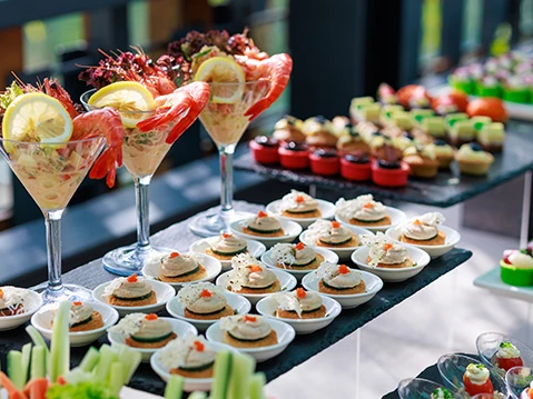 A fancy buffet with shrimp cocktails in tall glasses, assorted bite-sized canap&eacute;s on trays, and colorful desserts in the background.