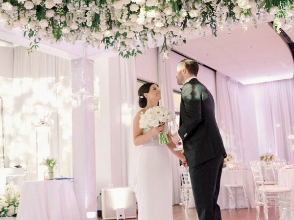 A couple in a wedding dress and suit admire each other under hanging blossoms in a softly lit, elegant ceremony space.