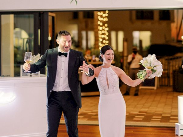 A newlywed couple enters a reception hall, smiling and dancing with bouquets, the groom in a tux and the bride in a white gown, celebrating joyfully.