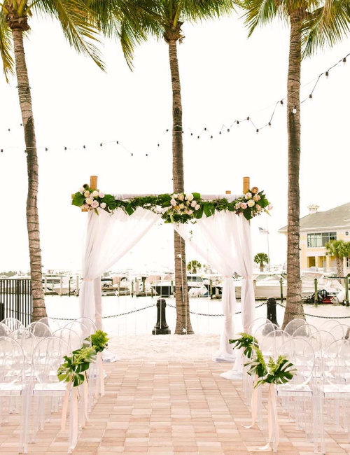 A tropical outdoor wedding setup with a white draped arch, string lights, palm trees, and clear acrylic chairs along a brick aisle by the water, decorated with greenery and flowers.