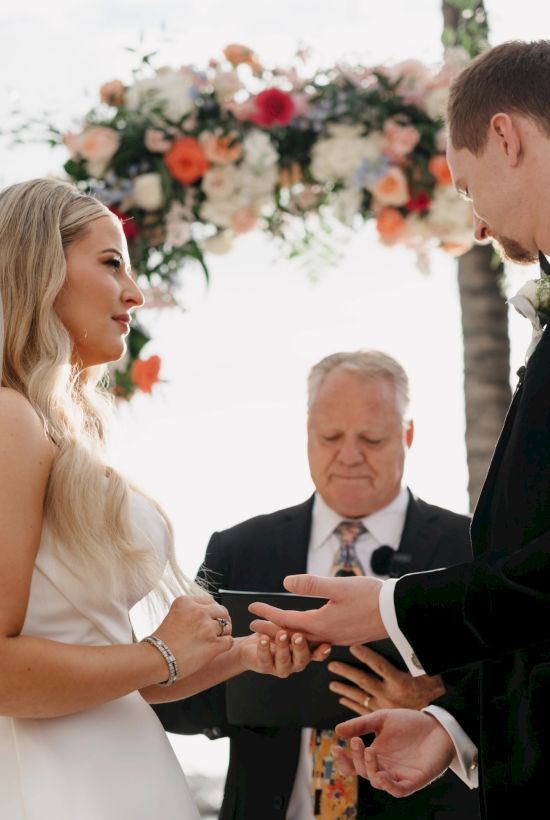 A bride and groom exchange rings under a floral arch at their outdoor wedding, with a officiant watching the ceremony.