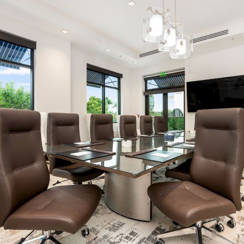 A modern office meeting room with glass windows, brown swivel chairs, a central conference table, and a wall-mounted TV.