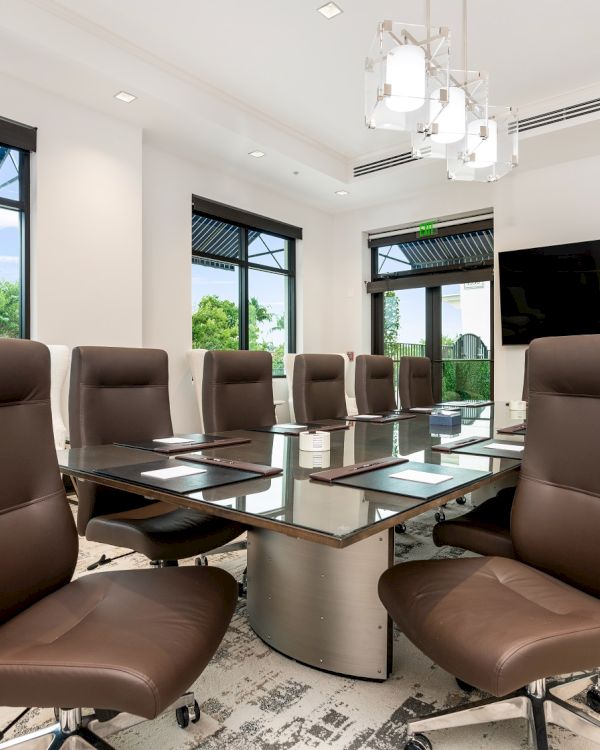 A modern office meeting room with glass windows, brown swivel chairs, a central conference table, and a wall-mounted TV.