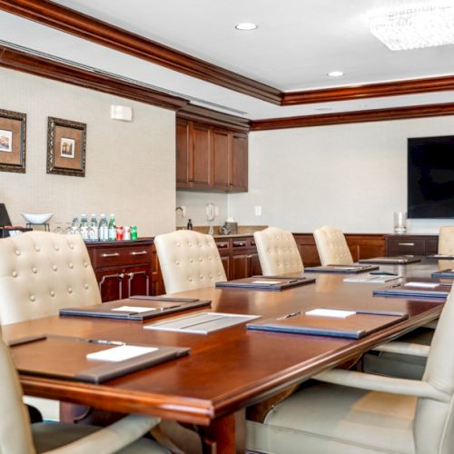 A formal conference room with a long wooden table, white leather chairs, framed pictures, and a large flat-screen TV.