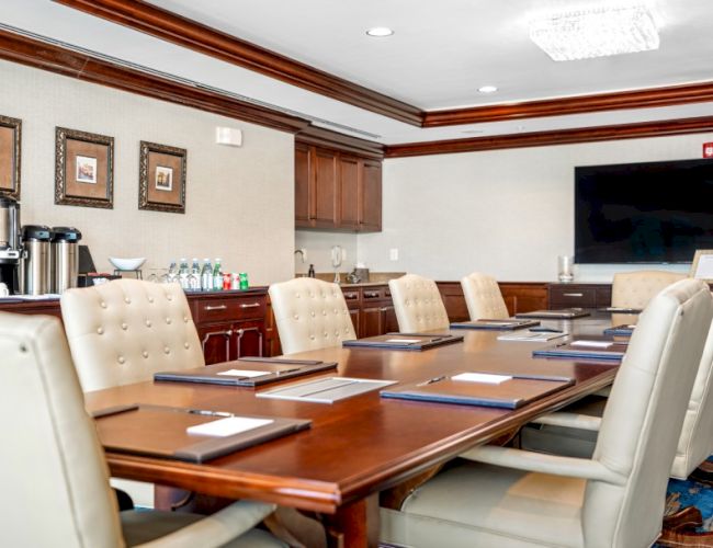 A formal conference room with a long wooden table, white leather chairs, framed pictures, and a large flat-screen TV.