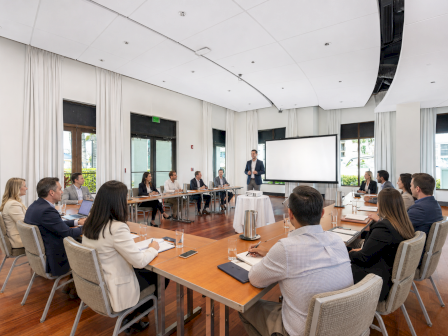 A business presentation in a modern conference room where a group of professionals sits around a long table, listening.