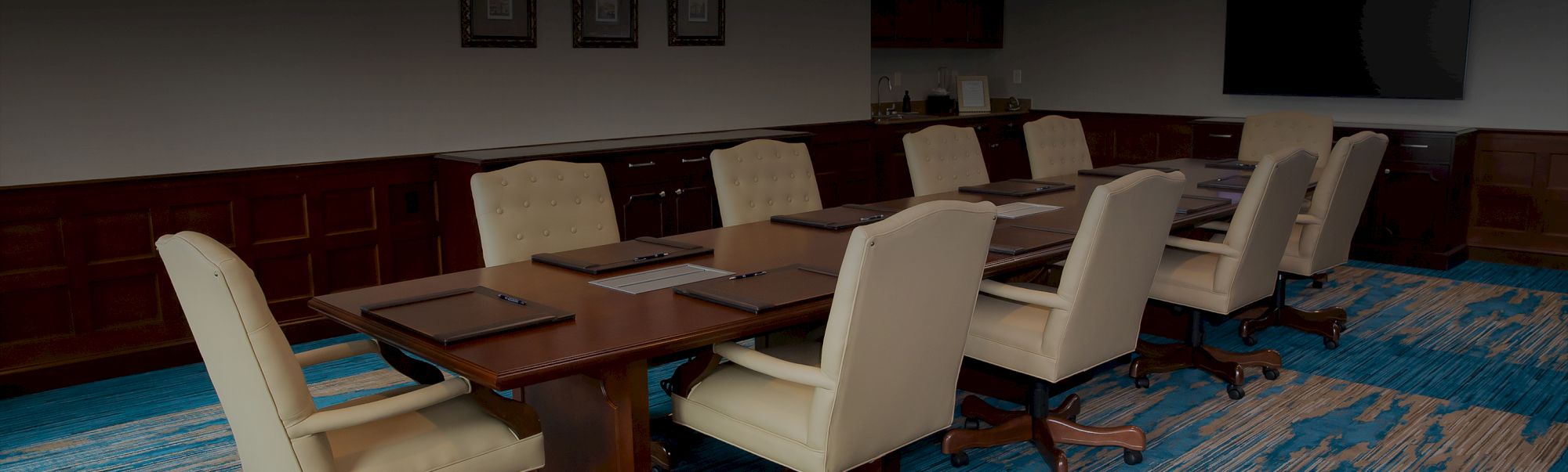 A long conference table with beige cushioned chairs around it, set in a formal meeting room with wood paneling and blue carpet.
