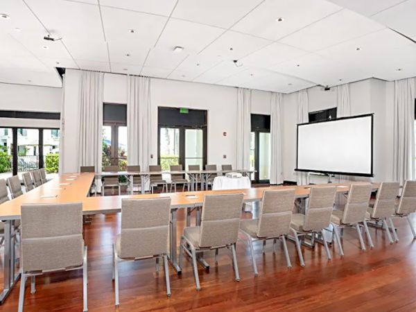 A bright conference room with a long wooden table, beige chairs, white walls, a projector screen, and large windows.