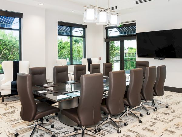 A modern conference room with a long glass table, brown rolling chairs, large windows, a wall-mounted TV, and a light patterned carpet.