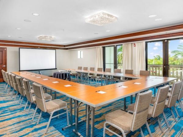 A bright, modern conference room with a large U-shaped table, many gray chairs, white walls, blue patterned carpet, and a projector screen at one end.