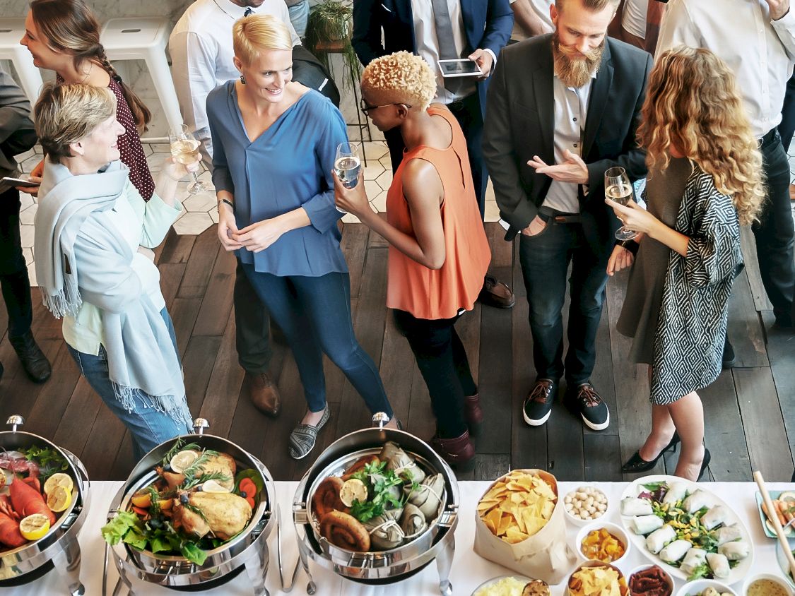 A group of professionals mingle at a catered event, chatting and sipping drinks around a buffet spread of food.