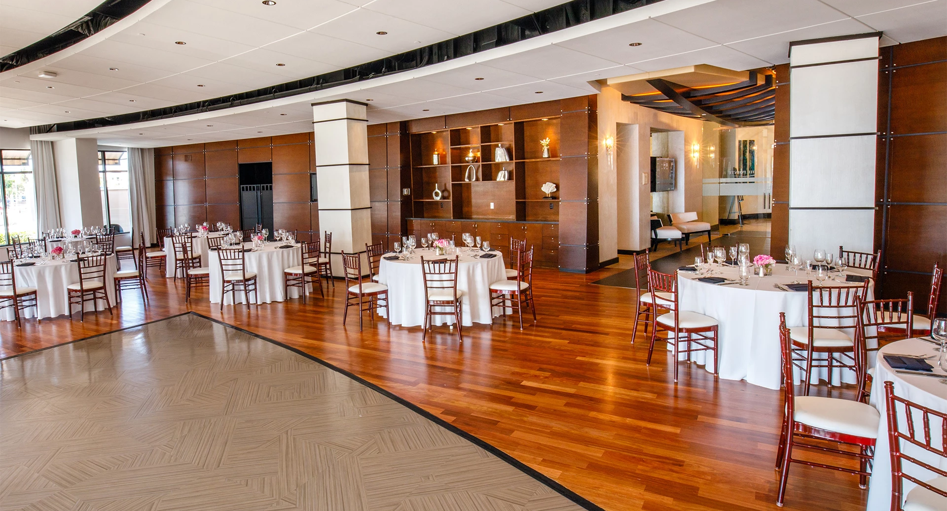 A bright banquet hall with round tables dressed in white, wooden chairs, warm wood paneling, and soft ceiling lighting.