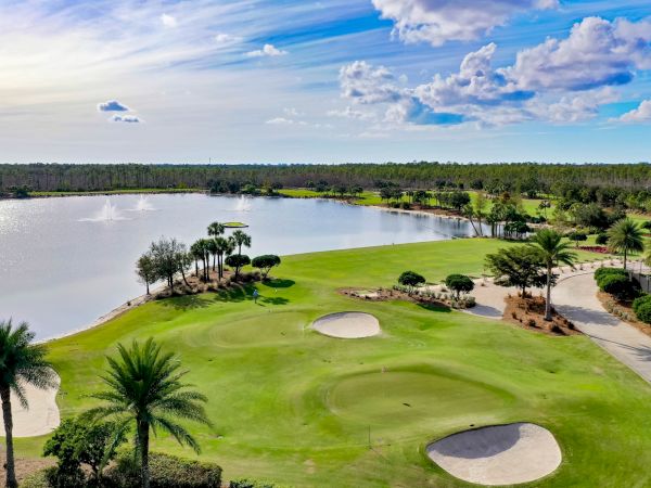 A sunny golf course beside a calm lake with fountains, lush greens, palm trees, and golfers under blue skies with clouds.