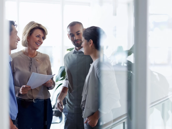 A diverse group of professionals in a modern office, sharing a conversation near a glass wall, with papers in hand and smiles.