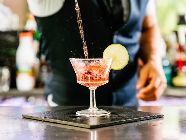 A bartender hands over a pink cocktail with a lime wedge on a drink glass, placed on a bar counter with a blurred busy background.