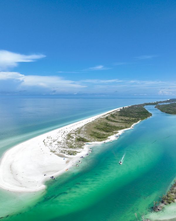 Aerial view of a tropical beach with turquoise water, boats, and a curving lagoon under a blue sky.