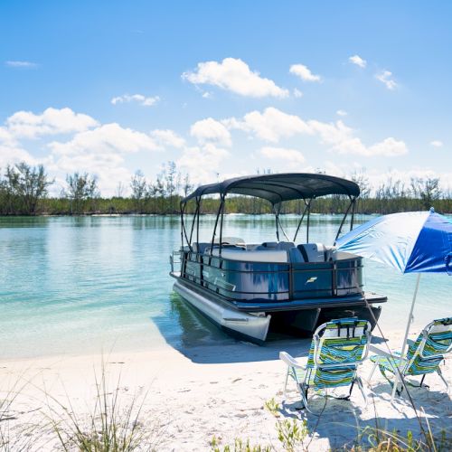 A pontoon boat is moored on a sunny lakeshore beside two striped chairs and a blue-and-white beach tent.