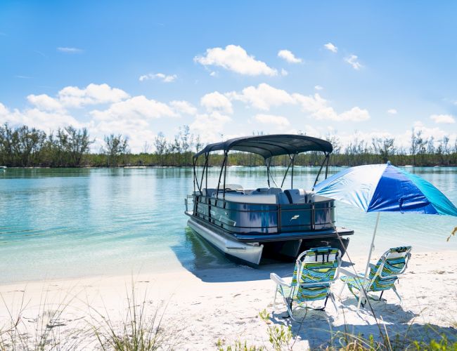 A pontoon boat is moored on a sunny lakeshore beside two striped chairs and a blue-and-white beach tent.