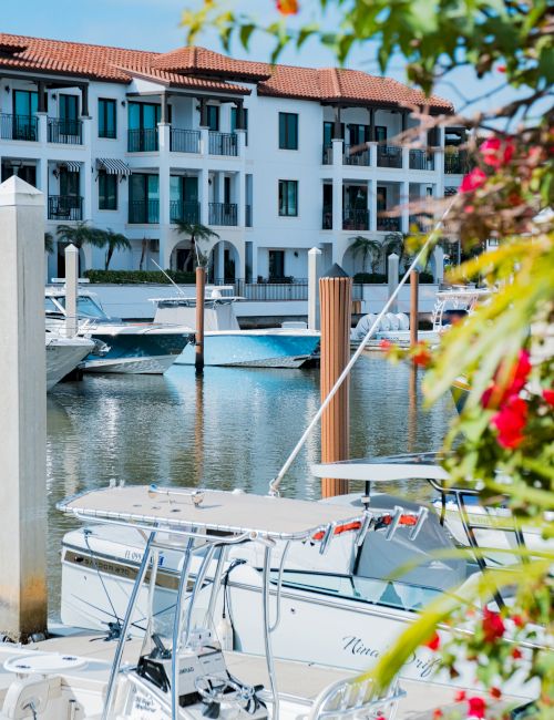 A marina scene with yachts docked in front of a white, terracotta-roofed apartment building, framed by colorful foliage.