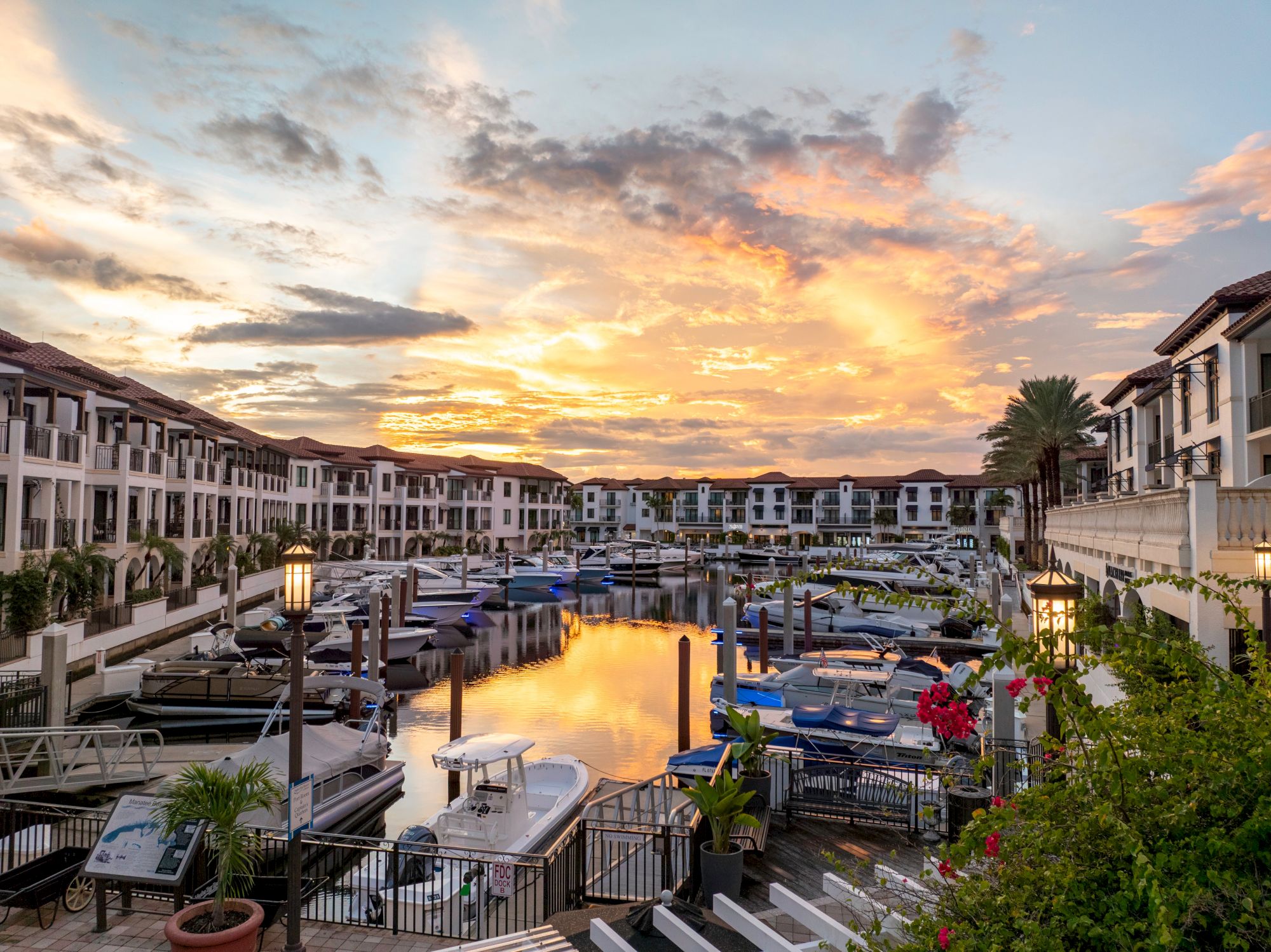 A tranquil resort lagoon at sunset, lined with boats, palm trees, and pastel-hued buildings reflecting in calm waters.