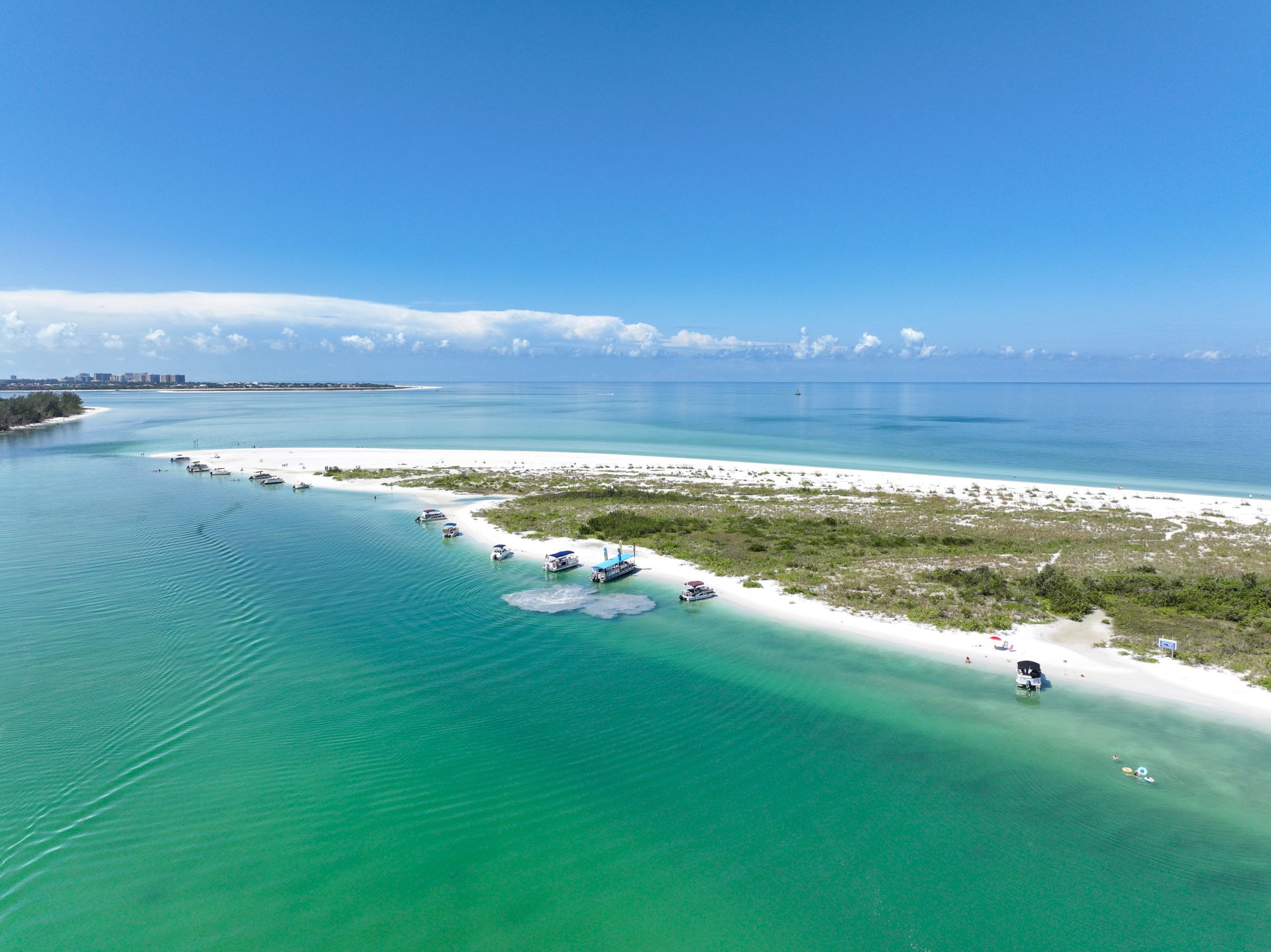 Aerial view of a tropical beach bay with clear turquoise water, sandy shoreline, and anchored boats under a blue sky.