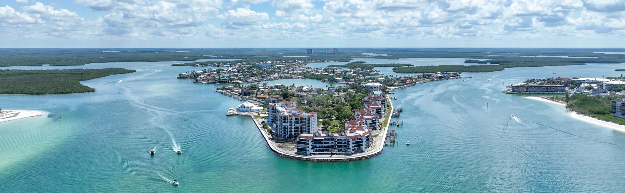 A small coastal island community with modern buildings, surrounded by turquoise water, boats, and canals under a partly cloudy sky.