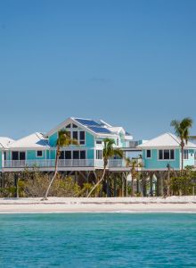 A turquoise beachfront house on stilts with blue-gray siding, palm trees, and a white sandy shore by calm turquoise waters.