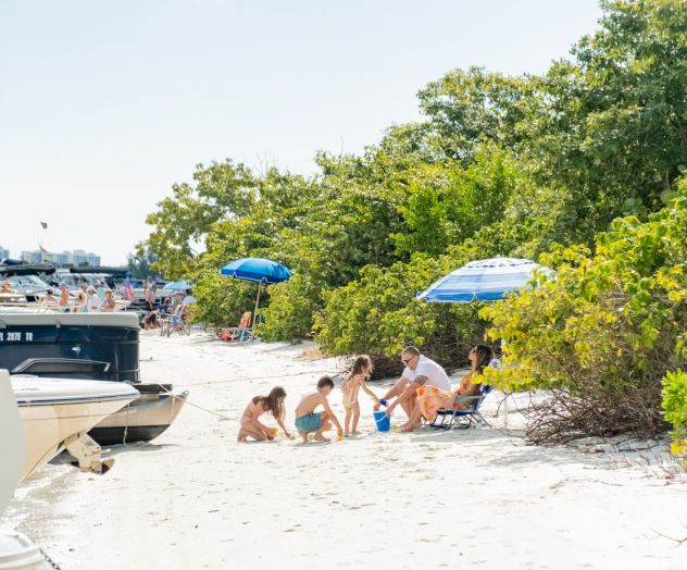 People relaxing on a sunny beach with umbrellas and boats, some kids building in the sand near green shrubs by the water.
