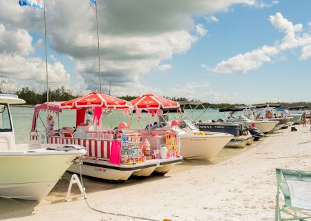 Colorful beach boats with pink-striped canopies line a sunny sandy shore, flags flutter overhead as calm water and buoys sparkle in the distance.