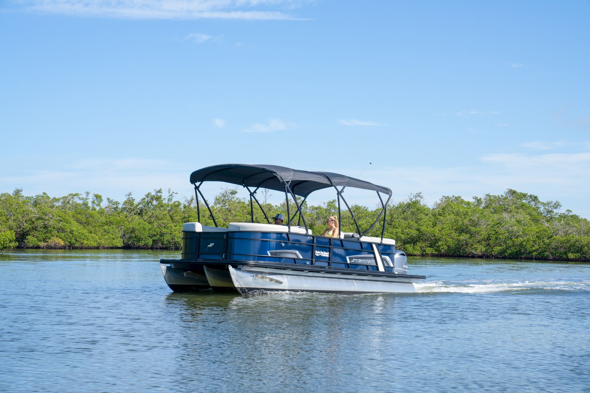 A blue pontoon boat with two people cruises on a calm lake under a bright blue sky.