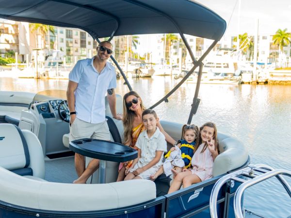 A family of five sits on a pontoon boat docked at a marina, with a man steering and a woman with a child smiling near the front.