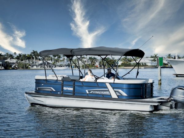 A blue pontoon boat with a canopy sails on calm water near a marina, passengers seated, clear sky and palm trees in the background.